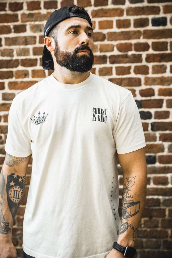 A bearded man in the 13th Christ is King Premium Tee - Ivory and a backwards black cap stands before a brick wall, displaying tattoos on both arms while looking slightly to the side.