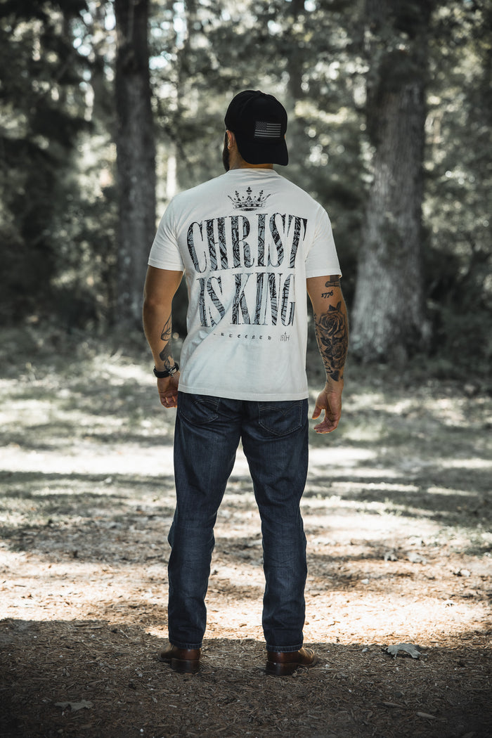 A man stands outdoors with his back turned, wearing the 13th Christ is King Premium Tee - Ivory, paired with jeans, a cap, and a watch in a sunlit wooded area.