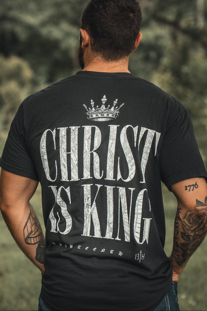 A man stands outdoors, his back to the camera, wearing the 13th Christ is King Premium Tee—black with bold white "Christ Is King" text and a crown graphic. His arms are covered in tattoos.