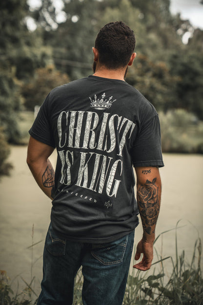 A man stands outdoors in the 13th Christ is King Premium Tee, featuring bold "CHRIST IS KING" text with a crown graphic. His tattooed arms and the lush greenery complete this striking tribute to faith.