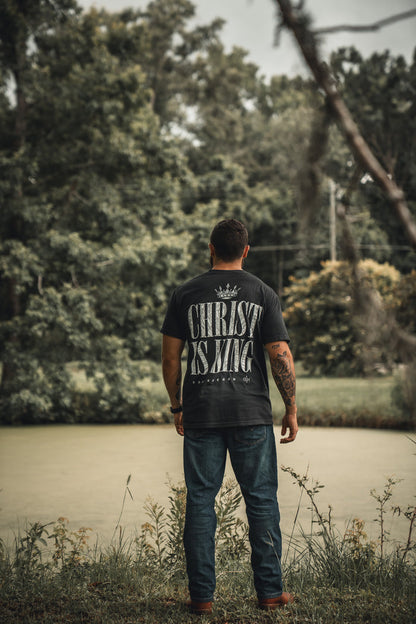 A man stands by a pond on an overcast day, surrounded by trees, wearing 13th's Christ is King Premium Tee—black with bold white text—and jeans.