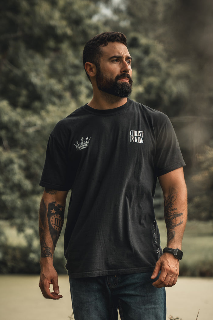 A bearded man with tattoos stands outdoors in jeans and the 13th Christ is King Premium Tee. Trees blur in the background, drawing focus to his bold expression of faith.