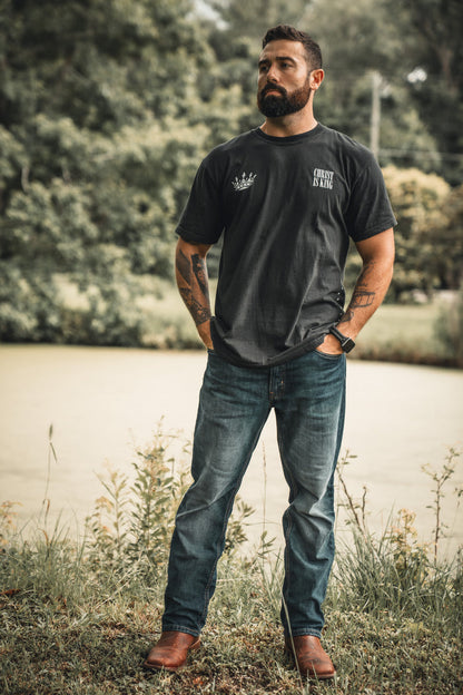 A bearded, tattooed man stands confidently by a pond amid trees, wearing the 13th Christ is King Premium Tee with blue jeans and brown boots, embodying serenity and strength in nature.
