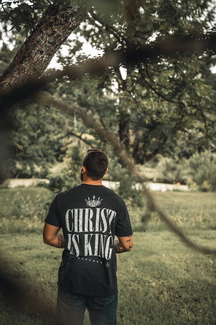 A man stands outdoors under a tree, wearing the 13th Christ is King Premium Tee—a black shirt with bold white “Christ is King” text and a crown graphic—proudly expressing his faith against a lush green backdrop.