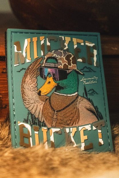 The Cool Duck PVC Patch by Mucker Bucket is a 4x4 patch featuring a duck wearing sunglasses and a camo hat, with "MUCKER BUCKET" in bold letters.