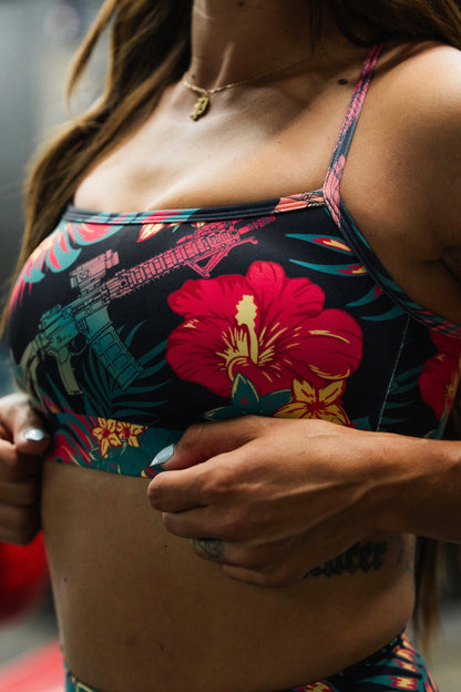 A woman with long hair, tattoos, and a small necklace adjusts the band of the Tango Charlie Apparel Cross Back Sports Bra | Tropical Assassin, featuring a bold gun graphic and vibrant tropical colors.