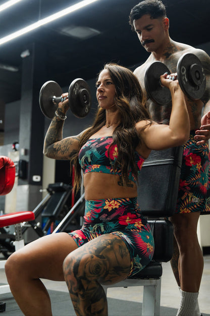 A woman wearing the Cross Back Sports Bra | Tropical Assassin by Tango Charlie Apparel lifts dumbbells overhead in a gym, seated with a man supporting her. Both have visible tattoos and focused expressions.