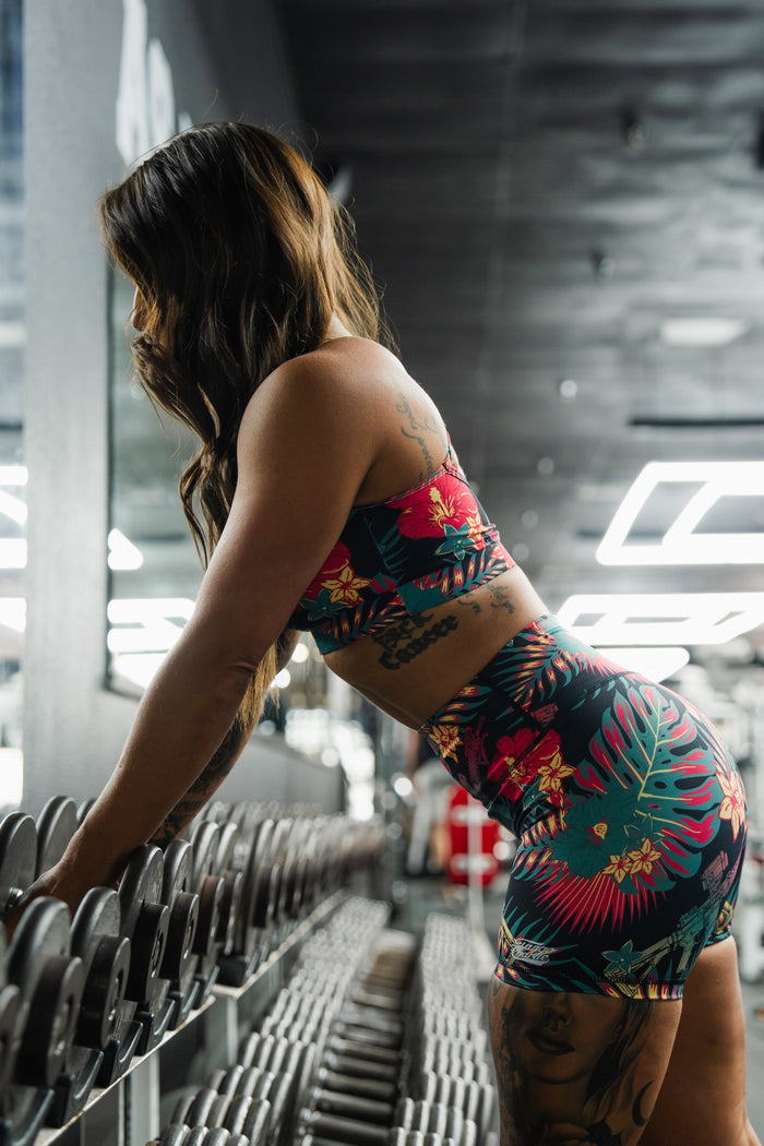 A woman in a vibrant Cross Back Sports Bra | Tropical Assassin by Tango Charlie Apparel leans over a rack of dumbbells at the gym, her side profile and the gym equipment visible as she prepares for strength training.