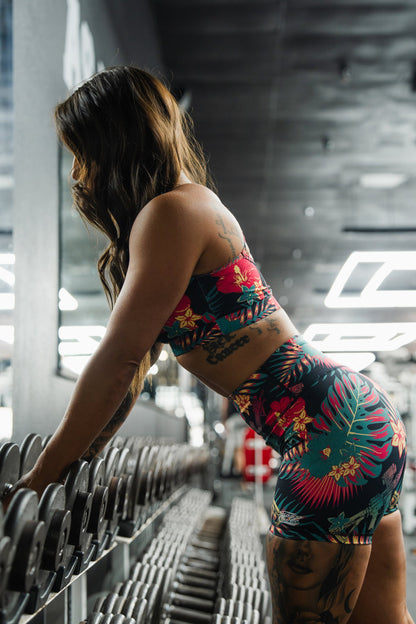 A woman in a vibrant Cross Back Sports Bra | Tropical Assassin by Tango Charlie Apparel leans over a rack of dumbbells at the gym, her side profile and the gym equipment visible as she prepares for strength training.