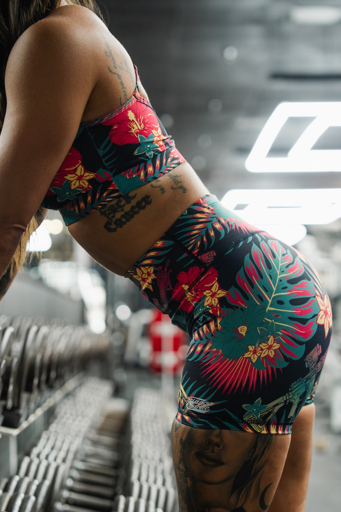 Wearing the Tango Charlie Apparel Cross Back Sports Bra | Tropical Assassin, a person with arm and leg tattoos leans near dumbbells in a gym. The gym background is blurred, highlighting the bra’s vibrant tropical pattern and adjustable cross-back straps.
