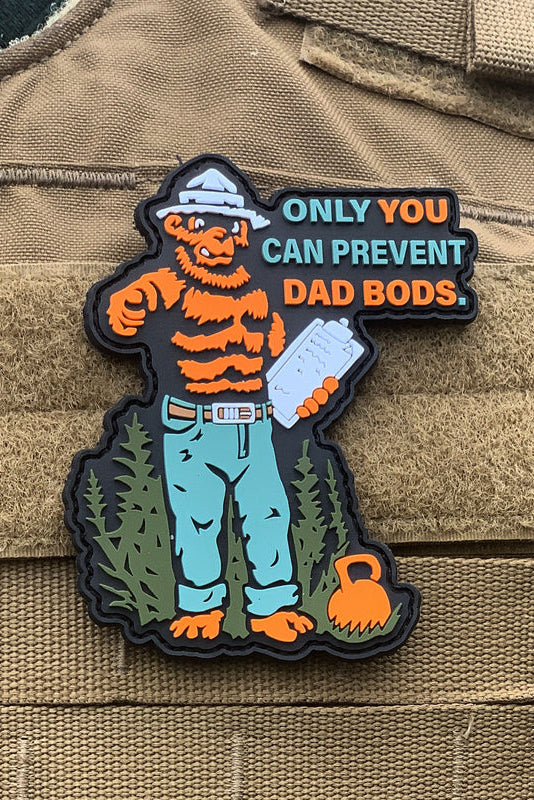 The Dad Bods Patch from Tango Charlie Apparel features a cartoon bear in jeans and a hat, lifting a dumbbell, with the message ONLY YOU CAN PREVENT DAD BODS. It’s designed with velcro for easy attachment to any tactical vest.