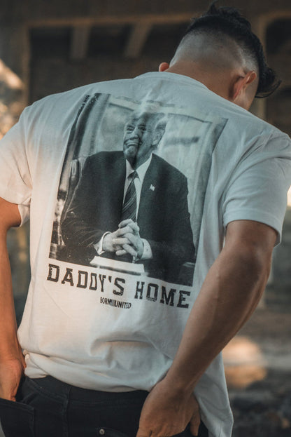 Outdoors and partially turned away, a person wears the Daddys Home - #47 - White tee by Born United Private Label, featuring a suited man with clasped hands and hinting at Trump’s second win.
