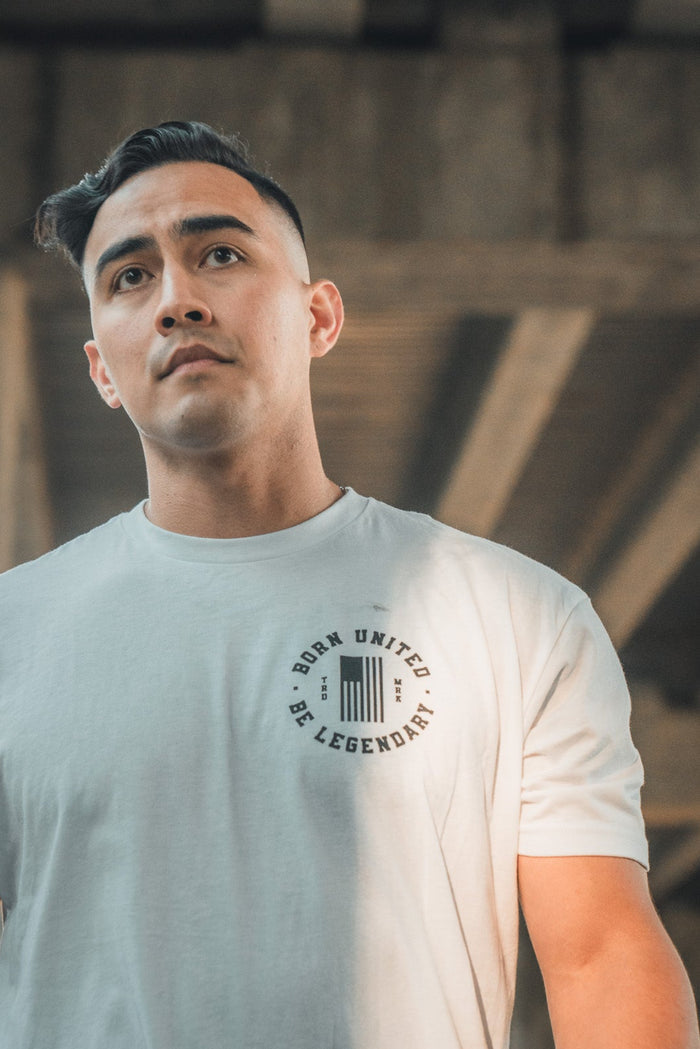 Under a large concrete structure, a person with short dark hair gazes upwards, wearing the Daddys Home - #47 - White premium tee from Born United Private Label. Sunlight and shadows dance across their face and shirt, showcasing the bold Born United Be Legendary text on their chest.