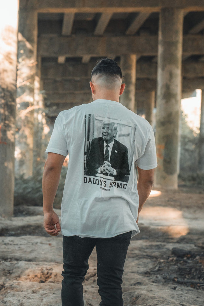 Wearing the exclusive pre-order Daddys Home - #47 - White tee by Born United Private Label, someone strolls beneath a wooden bridge outdoors. Sunlight filters through, casting playful shadows on the premium shirt featuring a graphic and the text DADDYS HOME.