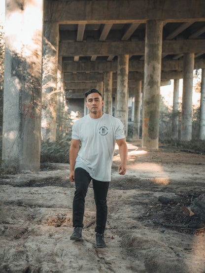 Wearing a Daddys Home - #47 - White tee from Born United Private Label and dark pants, a person strolls on a dirt path under a concrete bridge. Sunlight filters through columns, casting shadows amid the calm scene with trees and graffiti in the background.