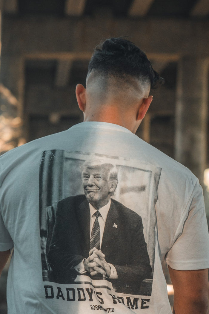 Under a dimly lit bridge, someone wears Born United Private Labels premium Daddys Home - #47 - White t-shirt featuring a suited figure with the text DADDYS HOME. This exclusive tee captures the mood of Trumps second victory. Pre-order yours now!.
