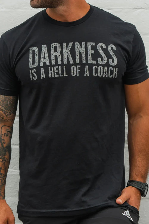 A man wears the Tango Charlie Apparel "Darkness is a Hell of a Coach" Tee, featuring bold distressed text and crafted from a comfyaf blend for stretch and all-day comfort.