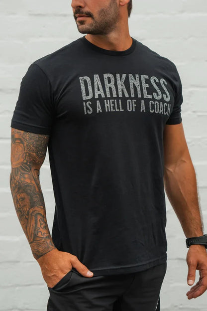 A man, hand in pocket, stands against a light gray background wearing the Tango Charlie Apparel "Darkness is a Hell of a Coach - Tee," a black T-shirt crafted from the comfyaf blend.