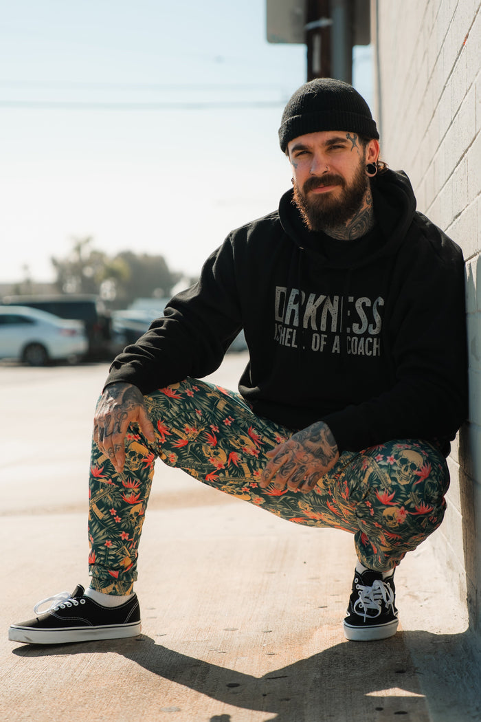 A bearded man in the Tango Charlie Apparel "Darkness is a Hell of a Coach Hoodie" and floral pants crouches outdoors against a wall on a sunny day, radiating triumph over darkness.