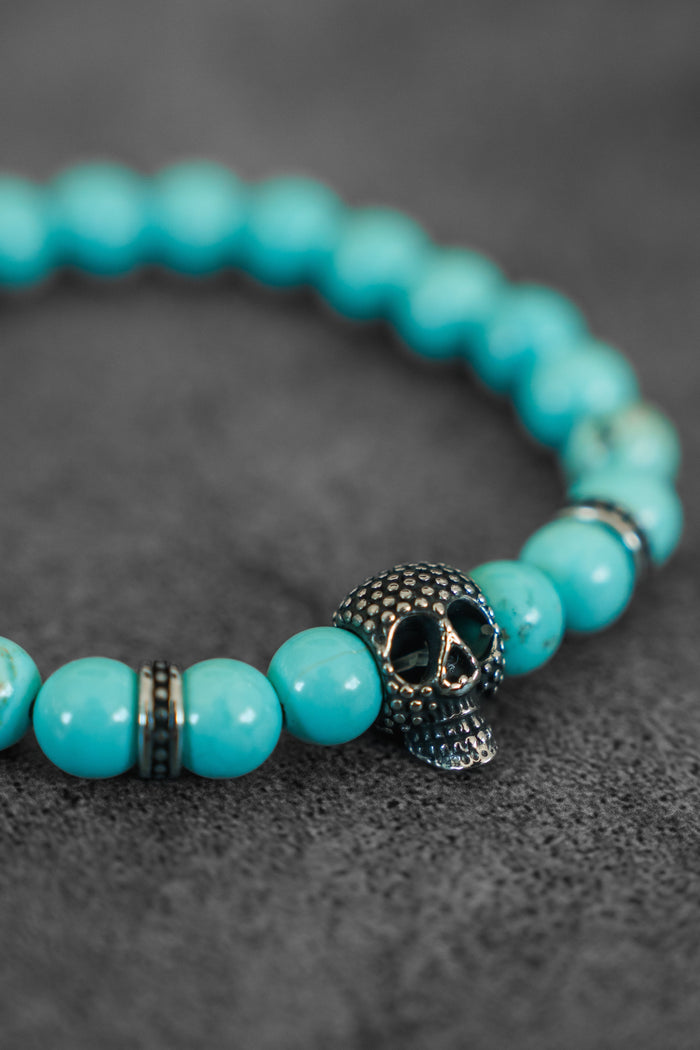 The Relentless Betrayal Day of the Dead Bead Bracelet showcases turquoise beads, a metallic skull charm, and metallic spacers, elegantly displayed on a textured dark gray surface.
