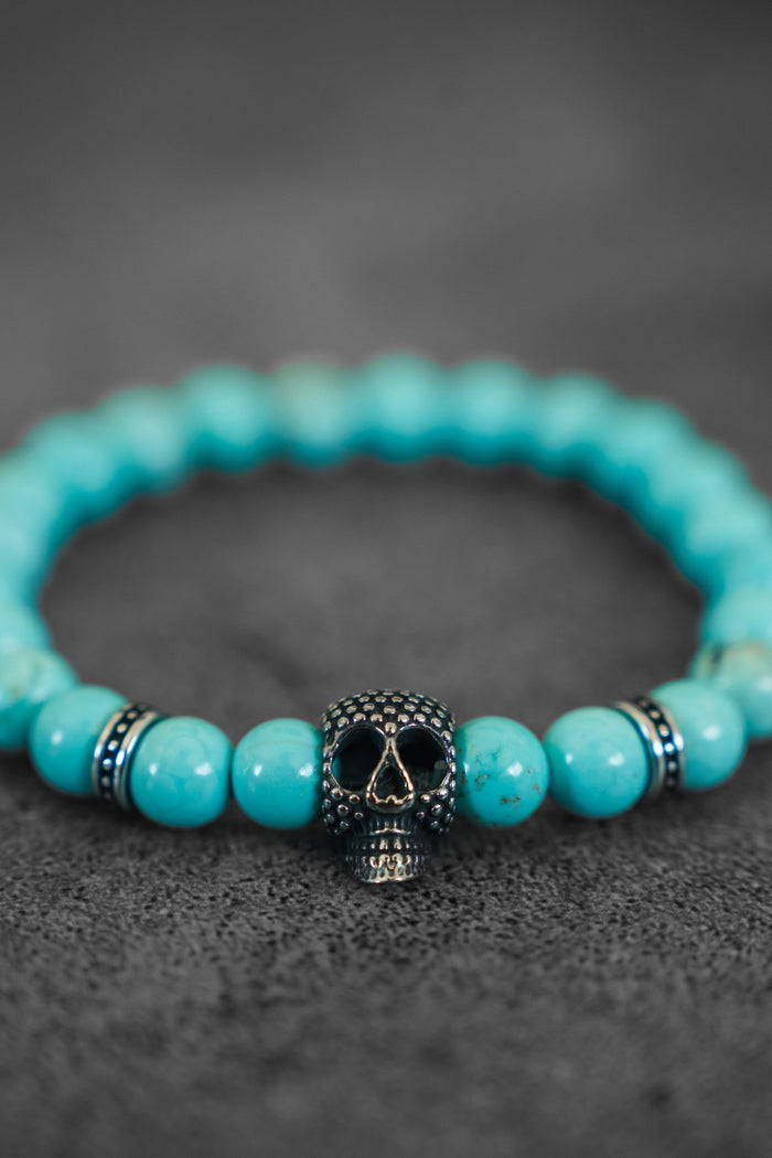 The Day of the Dead Bead Bracelet by Relentless Betrayal features turquoise beads and a metallic skull charm, making it a bold piece of skull jewelry displayed on a textured dark surface.