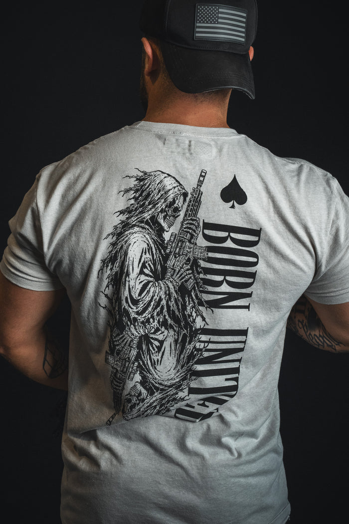 A person facing away from the camera wears the Born United Private Label’s Death Card tee in light gray, featuring a skeleton with a gun and “BORN UNLUCKY” emblazoned on the back, channeling a dark aesthetic.