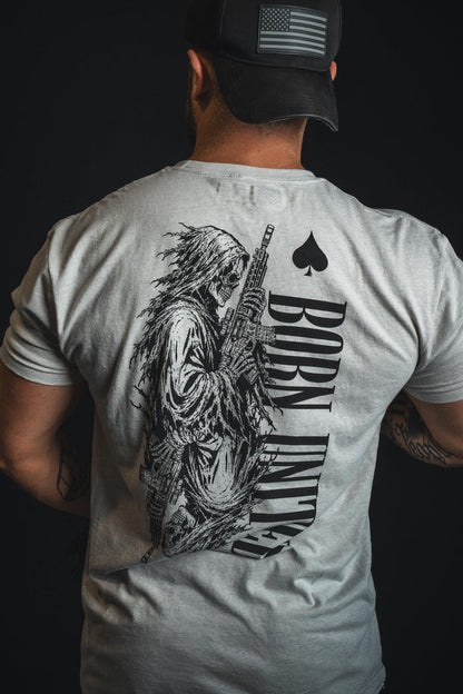A person facing away from the camera wears the Born United Private Label’s Death Card tee in light gray, featuring a skeleton with a gun and “BORN UNLUCKY” emblazoned on the back, channeling a dark aesthetic.