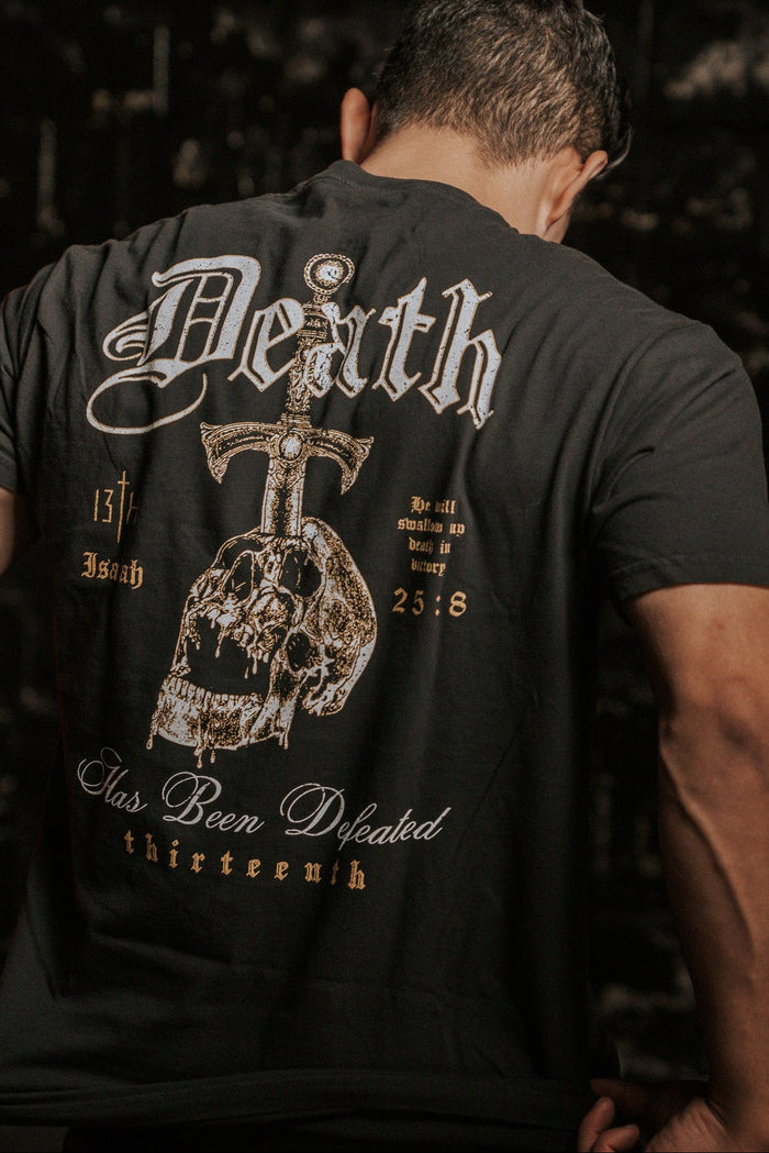 A man wears the 13th brand Death's Defeat Premium Tee, a black shirt with "Death Has Been Defeated" and a sword through a skull on the back, featuring Isaiah 25:8 for spiritual hope.