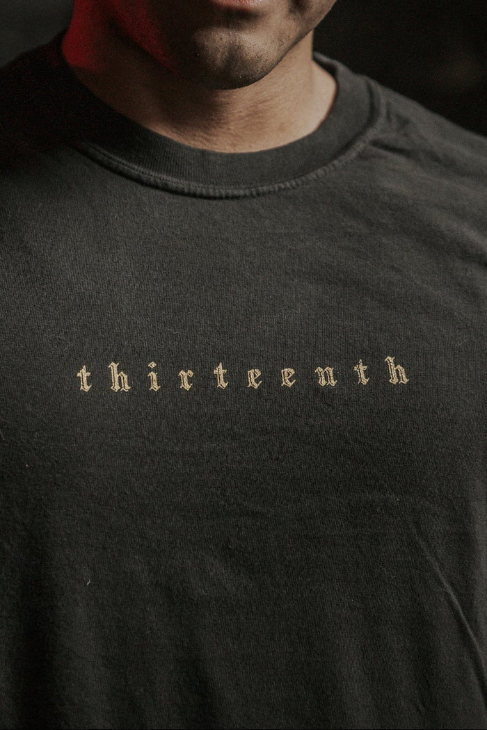Close-up of a person wearing the 13th brand Death's Defeat Premium Tee, featuring "thirteenth" in an old-style font across the chest. The shirt’s dark color subtly reflects spiritual hope; only the wearer’s lower face and neck are visible.