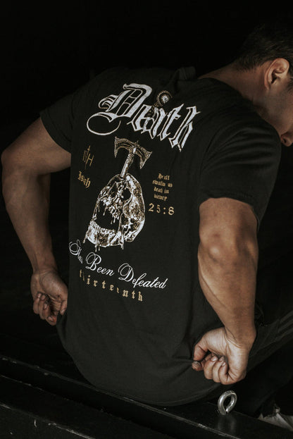 A person sits gripping a metal bar, seen from the back in low lighting, wearing the 13th Death's Defeat Premium Tee—a black graphic t-shirt featuring "Faith" and spiritual hope designs.