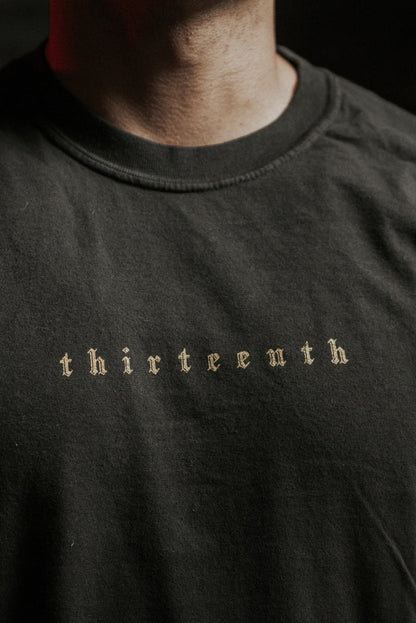 A close-up shows a person wearing the 13th Death's Defeat Premium Tee, featuring “thirteenth” in small, old English-style font. The dark look and background evoke Isaiah 25:8’s hope and victory over death.