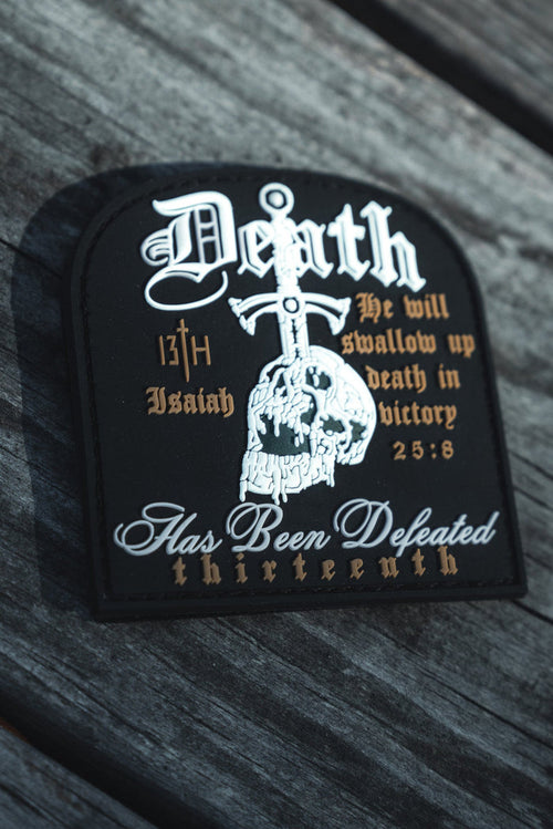 The Deaths Defeat PVC Patch by 13th lies on a wooden surface, showcasing gothic text and a skull. It includes biblical references like Death Has Been Defeated and Isaiah 25:8.