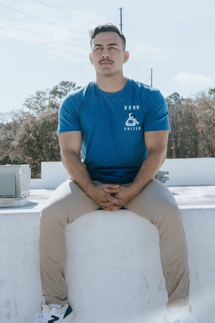 A disgruntled man sits on a white ledge outdoors, wearing a blue Disabled Vet t-shirt from Born United Private Label, beige pants, and New Balance sneakers. The background features trees and a blue sky with light clouds.