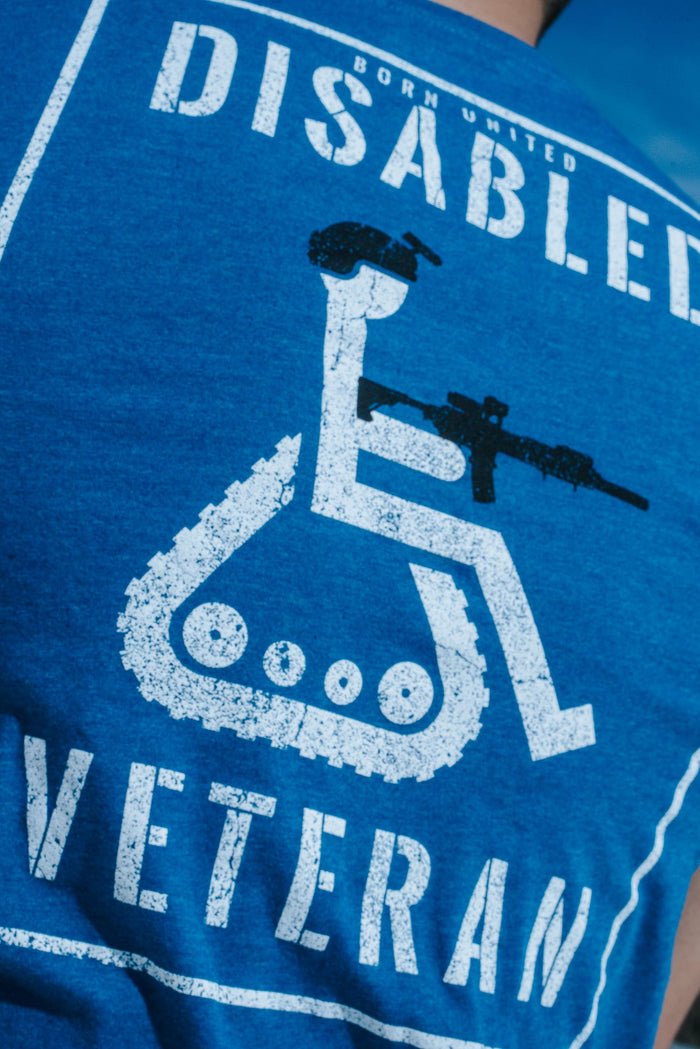 Back view of the Born United Private Label Disabled Vet - Blue T-shirt, featuring a wheelchair symbol with a helmet and rifle, along with Disabled Veteran text. The wearers disgruntled expression complements the outdoor setting against a tranquil blue sky.