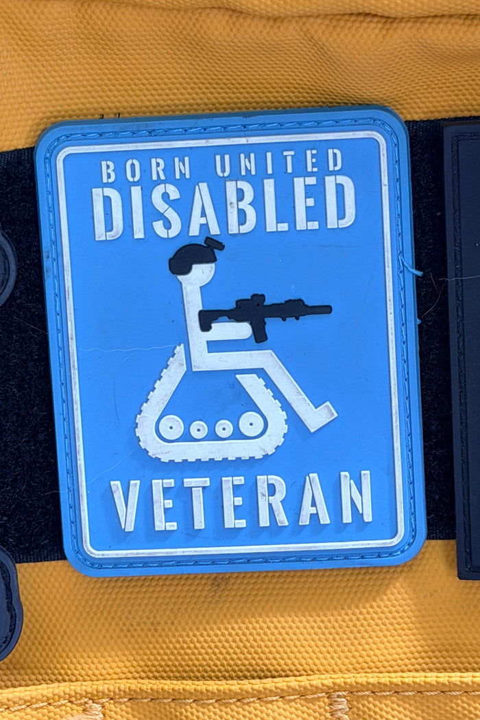 The Disabled Vet PVC Patch by Born United Private Label features a blue patch with a white wheelchair icon holding a rifle and bold "Born United Disabled Veteran" text, displayed against a yellow background.