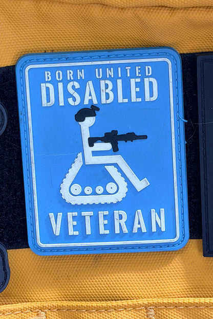 The Disabled Vet PVC Patch by Born United Private Label features a blue patch with a white wheelchair icon holding a rifle and bold "Born United Disabled Veteran" text, displayed against a yellow background.