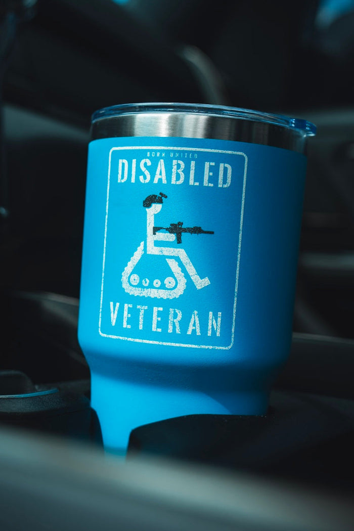 The Born United Private Label Disabled Vet Tumbler - 30oz, in blue stainless steel with a white helmet-and-rifle wheelchair graphic and text, is shown on a car’s center console.