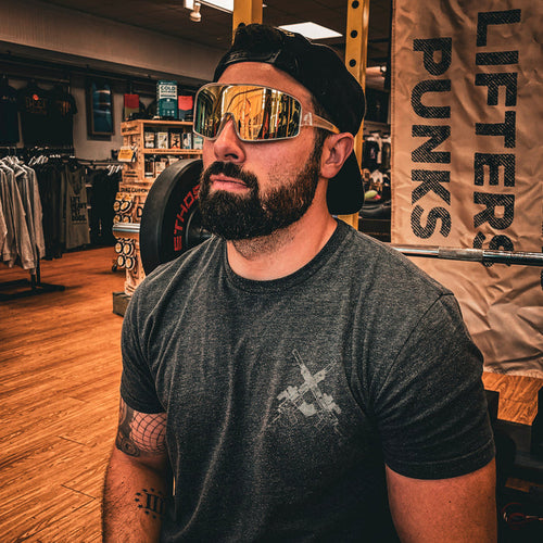 A bearded man in a gray t-shirt, backward cap, and Drippy Clear Sunglasses by Drippy Swimwear sits in a gym or fitness store with gym equipment and a LIFTERS PUNKS banner behind him.