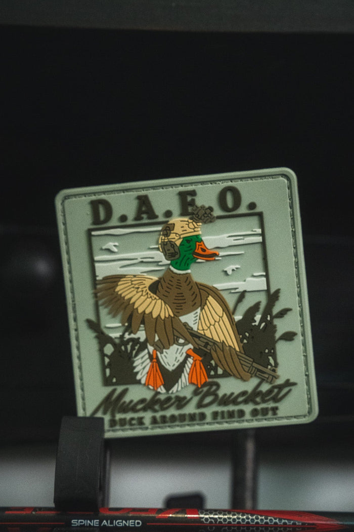An embroidered duck patch with the text 'D.A.F.O.' and 'Mucker Bucket Duck Around Find Out' displayed on a stand.