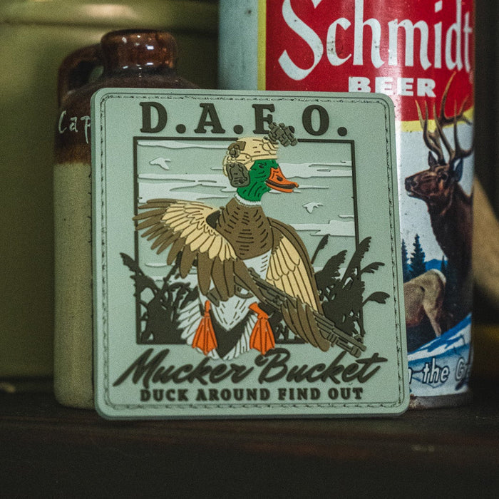 A Duck Around & Find Out PVC Patch by Mucker Bucket, measuring 4x4 inches and featuring an illustrated duck in military gear, is displayed in front of a Schmidt beer can.