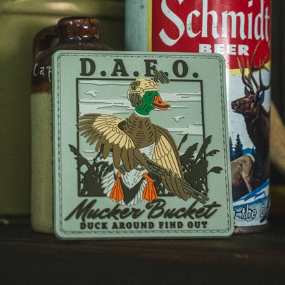 A Duck Around & Find Out PVC Patch by Mucker Bucket, measuring 4x4 inches and featuring an illustrated duck in military gear, is displayed in front of a Schmidt beer can.