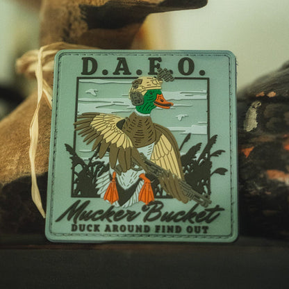 Show off your style with the Duck Around & Find Out PVC Patch by Mucker Bucket! Measuring 4x4 inches, this durable 3D PVC Velcro patch displays a cartoon duck in military gear with the bold "D.A.F.O. Mucker Bucket" and "Duck Around Find Out" text, all set against a detailed nature background—the perfect way to add personality and eye-catching flair to your gear.