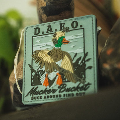 Introducing the Duck Around & Find Out PVC Patch by Mucker Bucket—a bold 4x4 Velcro patch showcasing a helmeted duck armed with a rifle and featuring the phrase "D.A.F.O. Mucker Bucket Duck Around Find Out" in striking 3D PVC detail.
