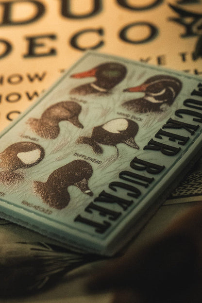 A “Duck DNA PVC Patch” by Mucker Bucket, featuring illustrations and names of five duck species, rests on a printed surface with plant shadows, resembling a detailed collectible 4x4 Velcro patch.