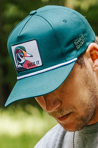 A man wears the Mucker Bucket Duck It - OG Green Wood Duck Bucket Hat, featuring a wood duck patch and "DUCK IT" text, white rope above the brim, Mossy Oak branding on the side, as he looks down.