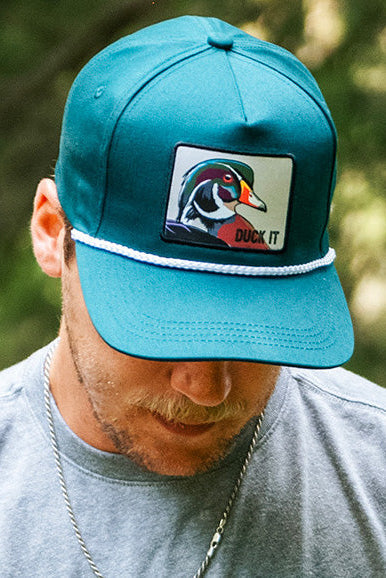 Wearing Mucker Bucket’s Duck It - OG Green Wood Duck Bucket Hat, a person in a gray shirt and silver chain looks down, standing before blurred greenery.