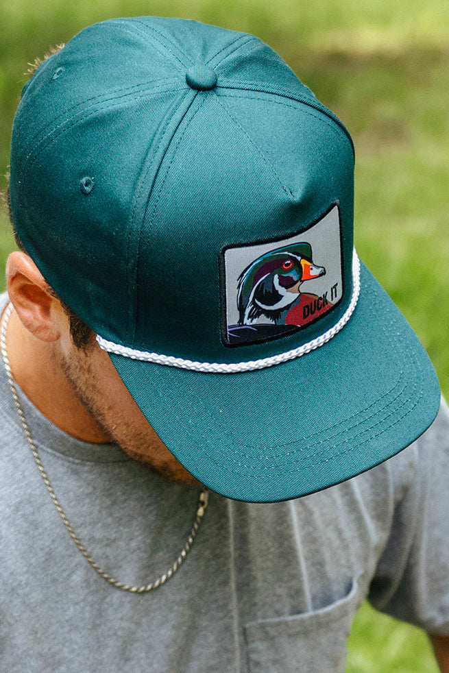 A person wearing the Mucker Bucket "Duck It - OG Green Wood Duck Bucket Hat" pairs it with a silver chain necklace and gray shirt, looking down in a grassy setting.