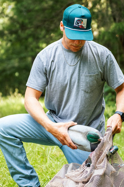 A man wearing a Mucker Bucket Duck It - OG Green Wood Duck Bucket Hat and gray t-shirt kneels on grass, holding a duck decoy and mesh bag, with trees in the background.