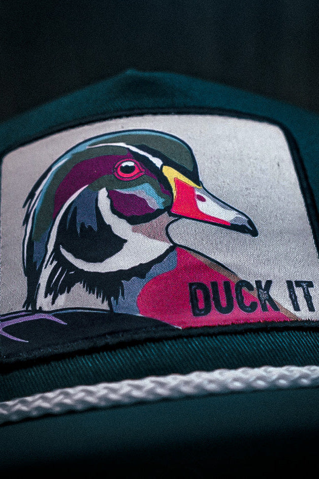 The Mucker Bucket Duck It - OG Green Wood Duck bucket hat features a handmade design with a high-quality embroidered patch of a colorful duck and DUCK IT, complemented by a white braided rope above the brim.