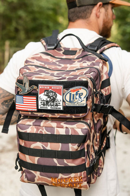 A man in a white shirt and cap carries the Mucker Bucket Duck It Limited Edition Tac-Pac - 25L tactical backpack, decorated with patches like an American flag, a duck, and "Life." Trees can be seen in the background.